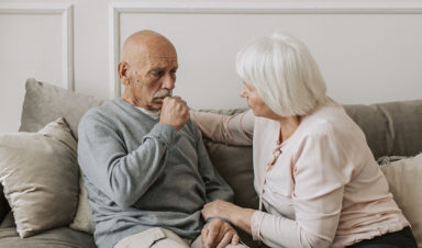 An older man with mesothelioma and an older woman sit on a couch as he coughs.