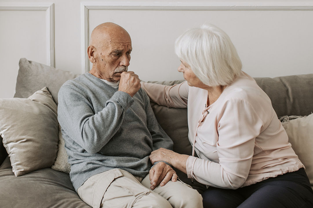 An older man with mesothelioma and an older woman sit on a couch as he coughs.
