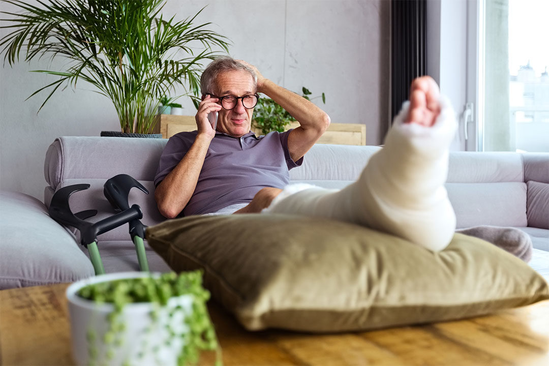 An older man with glasses sits on a couch with his foot elevated in a cast, and is on the phone with a lawyer to discuss a personal injury claim.
