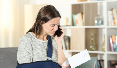 A woman wearing a sling over her right arm calls her insurance company on the phone while looking at a statement, with concerns they are acting in bad faith.