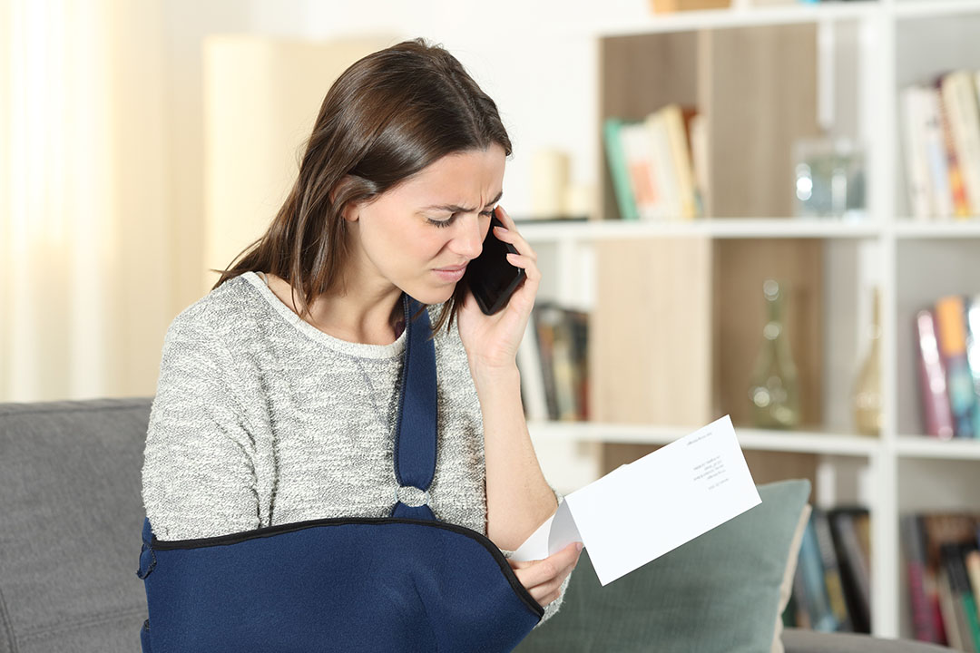 A woman wearing a sling over her right arm calls her insurance company on the phone while looking at a statement, with concerns they are acting in bad faith.