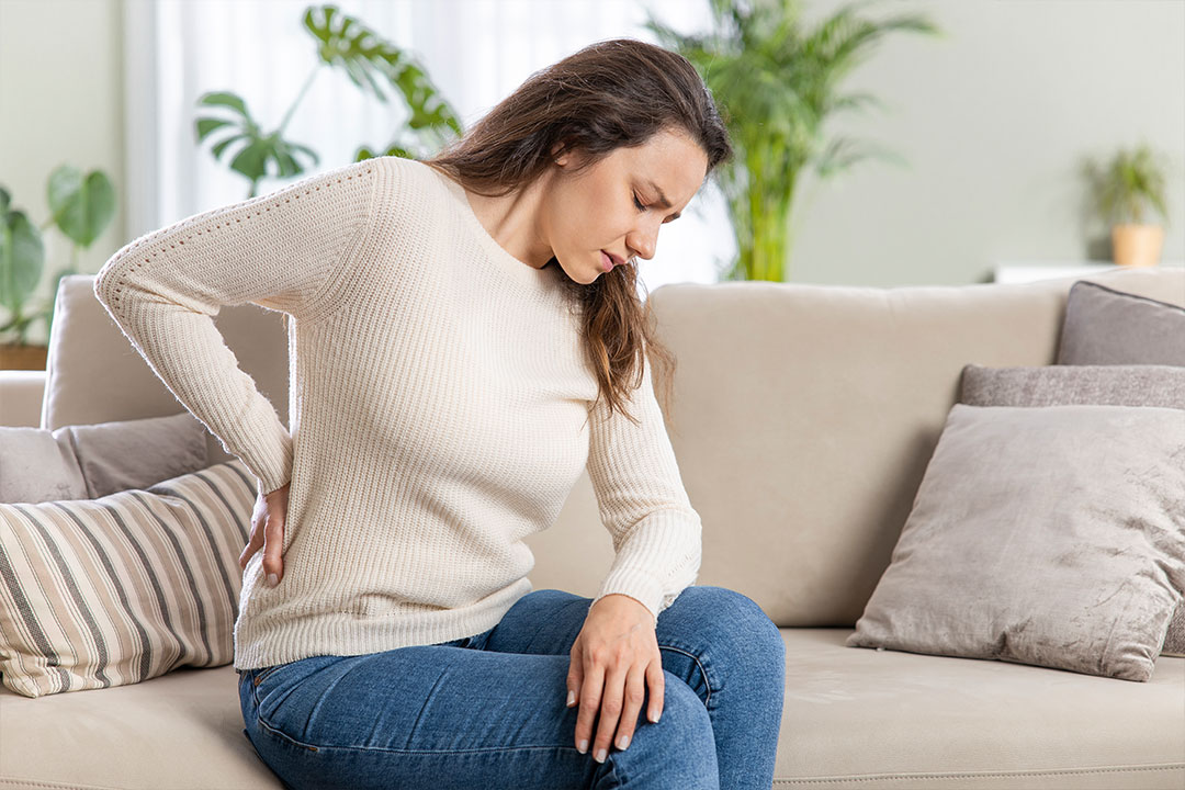 A woman sits on a couch and holds her back in pain, debating filing a personal injury claim.