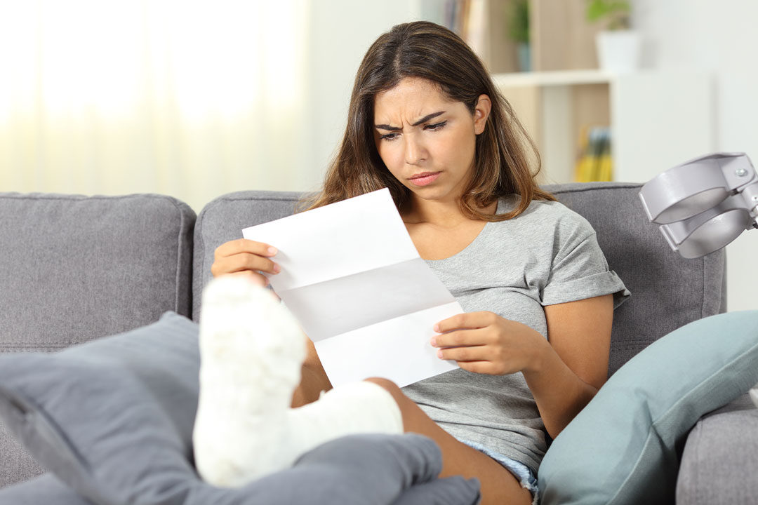 A woman sits on a couch and elevates her foot in a cast, reading a letter from her insurance company which is acting in bad faith.