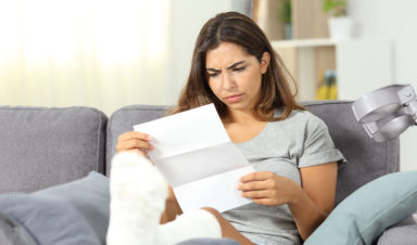 A woman sits on a couch and elevates her foot in a cast, reading a letter from her insurance company which is acting in bad faith.