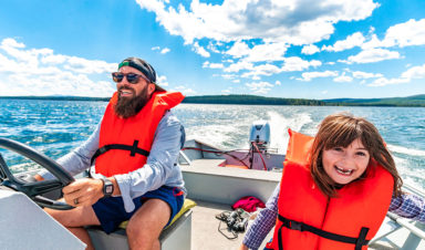 A father and daughter smile and laugh while driving a boat on a lake, each of them wearing a personal flotation device.