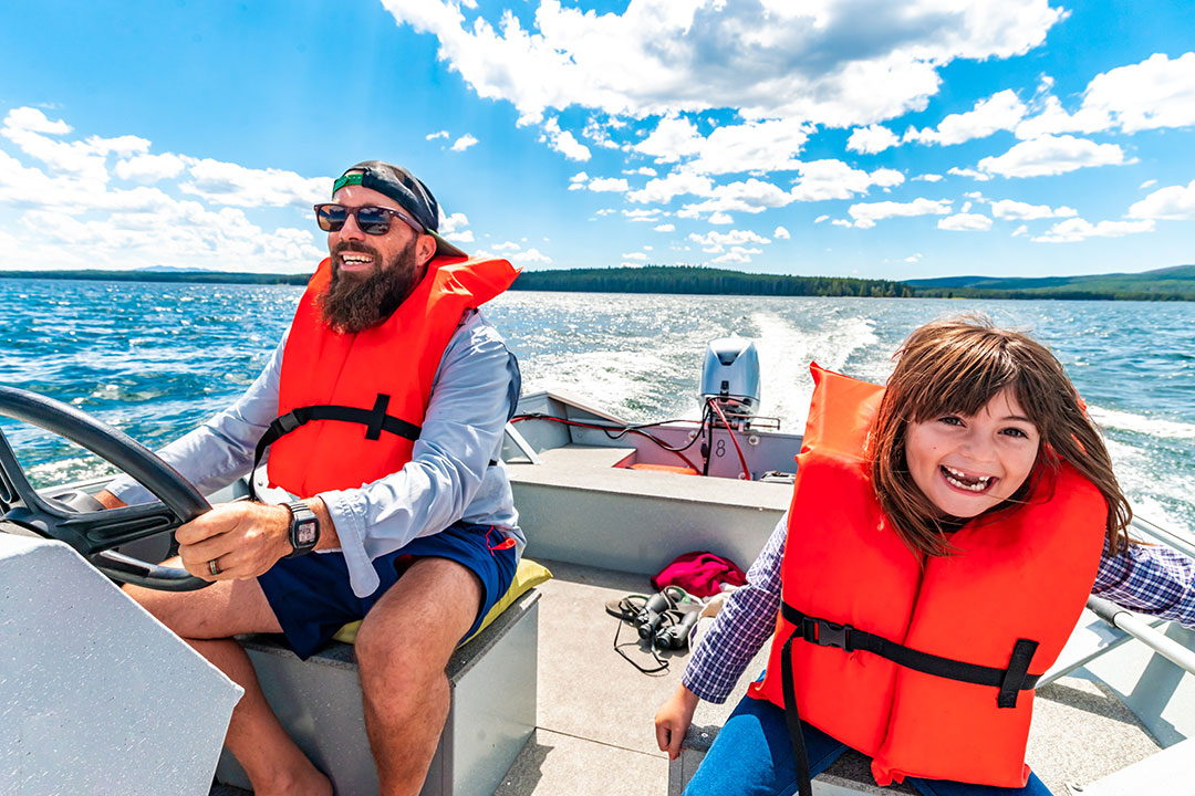 A father and daughter smile and laugh while driving a boat on a lake, each of them wearing a personal flotation device.