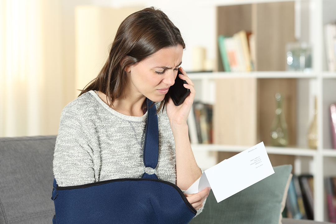A woman wearing a sling on her arm speaks to an unfair claims lawyer on the phone while looking at a bill in her hand