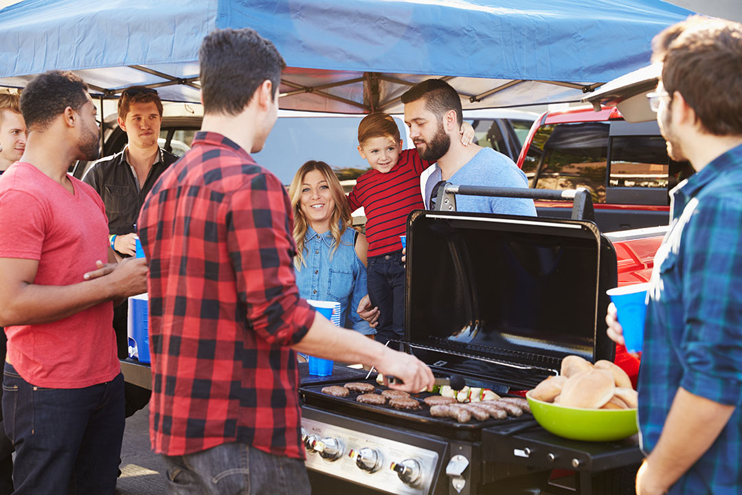 A group of young people and families gather around a grill in a tailgating parking lot before a football game.