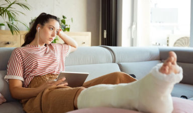 A young woman suffering from slip and fall injuries sits on her couch with a book and headphones in, with her leg in a cast and elevated on a pillow.