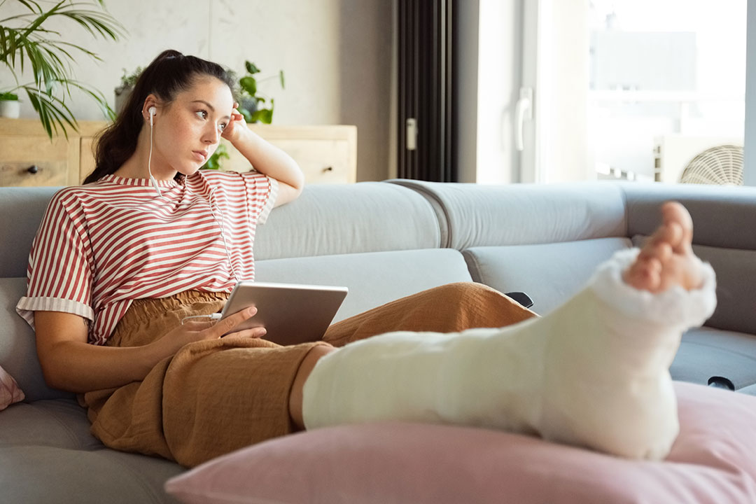 A young woman suffering from slip and fall injuries sits on her couch with a book and headphones in, with her leg in a cast and elevated on a pillow.