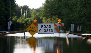 A "high water" sign and "road closed" sign indicate to drivers that flash flooding safety tips should be followed. The road is completely flooded.