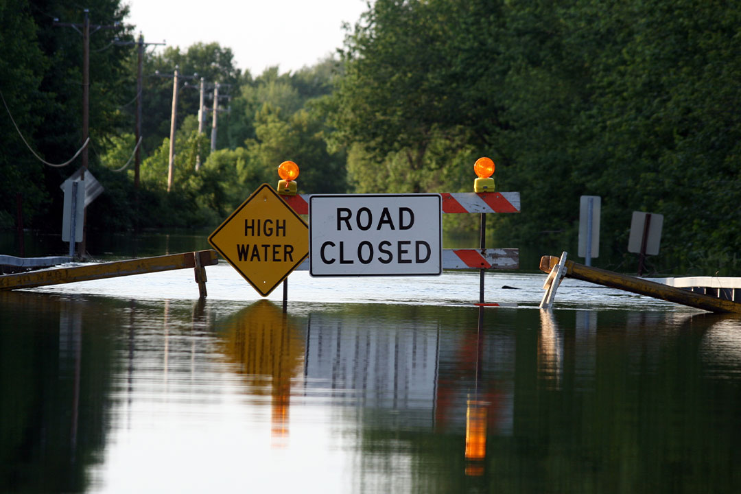 A "high water" sign and "road closed" sign indicate to drivers that flash flooding safety tips should be followed. The road is completely flooded.