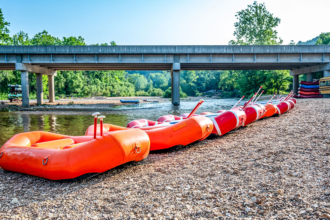 Six orange rafts are lined up on the bank of a river, prepped with oars in each.